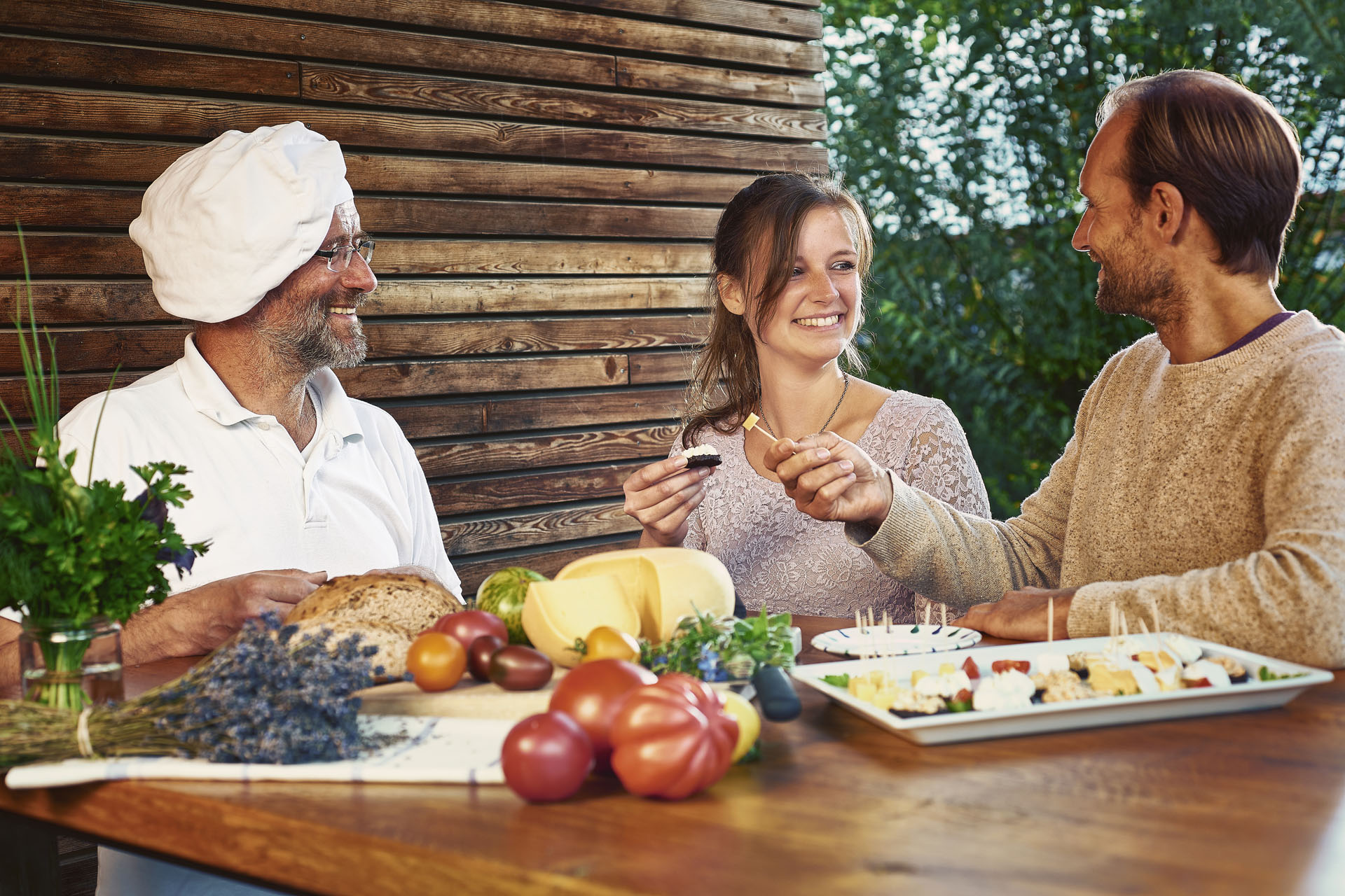 A couple and a cook on the left of them. The couple try small snacks from a plate next to him. Before that, a plate with bread, cheese and tomatoes.